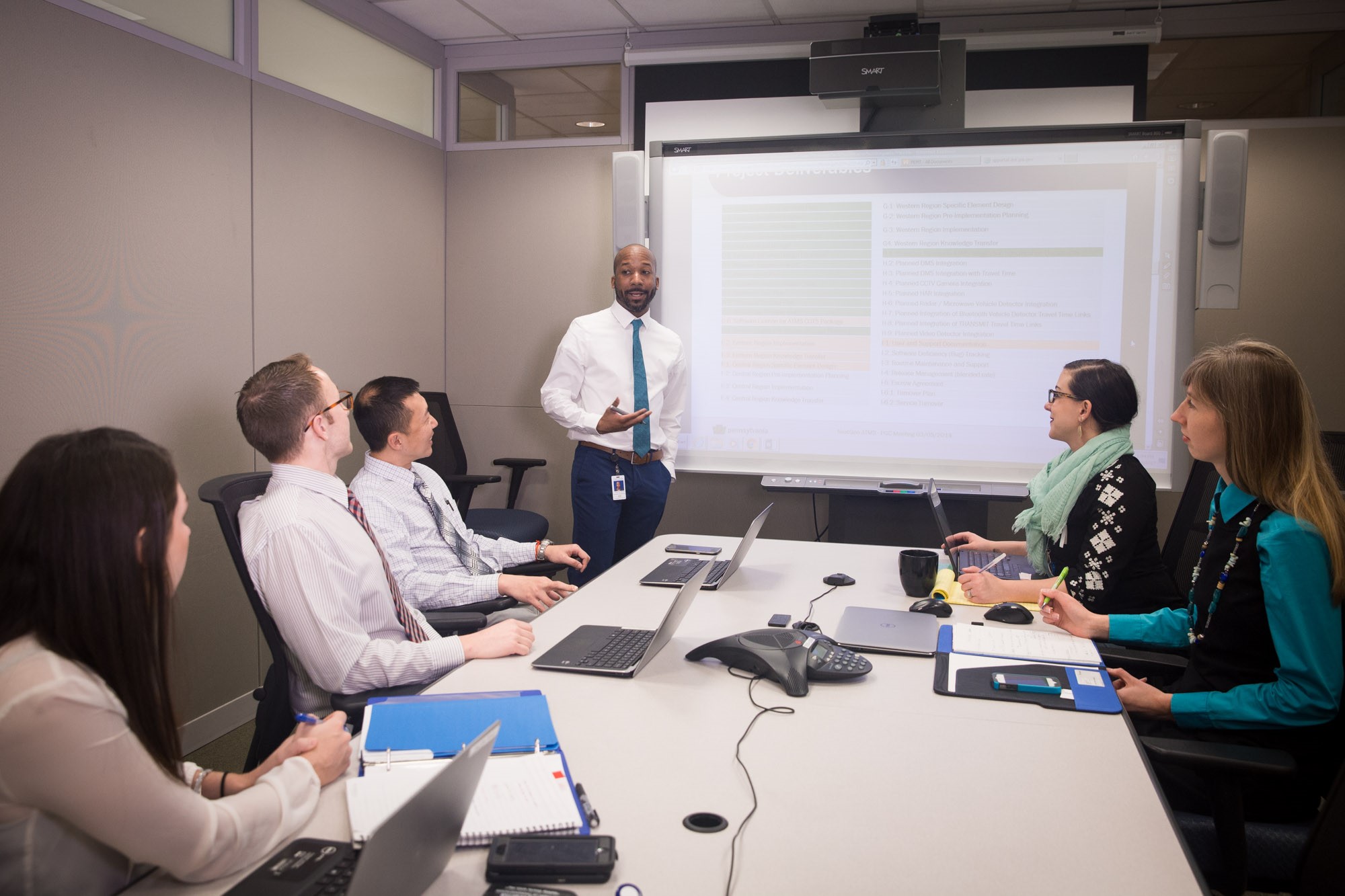 Employees in a conference room are listening to a presentation and are viewing information displayed on a Smart board